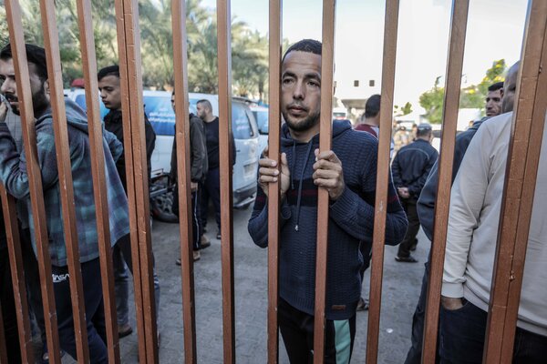 A man holds his head against the metal bars of a fence as others stand near him. There are cars in the background.